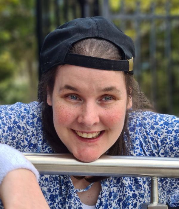 Jocelyn is resting her head on a metal bar. She is wearing a black cap, backwards, and a blue top. She is smiling toward the camera.