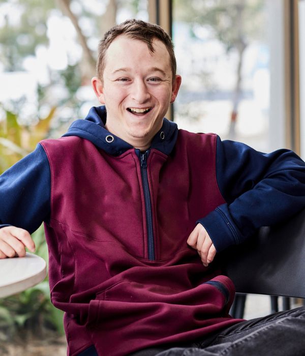 Luke is sitting on a chair with one arm resting on the table, and one leg over the other. He is wearing a maroon jumper and is smiling at the camera