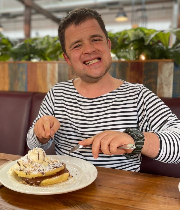 Daniel is wearing a black and white striped jumper, has short brown hair and is smiling at the camera. He is sitting at a table, eating a plate of pancakes.